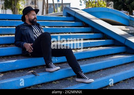 Lateinamerikanischer erwachsener Mann mit Bart und Hut, der entspannt auf der Treppe eines Platzes sitzt. Hipster- und Urlaubskonzept Stockfoto