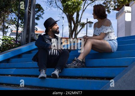 Erwachsener Mann mit Bart und Hut und junge lateinamerikanische Frau sprechen entspannt auf der Treppe eines Platzes. Tagungs- und Urlaubskonzept Stockfoto