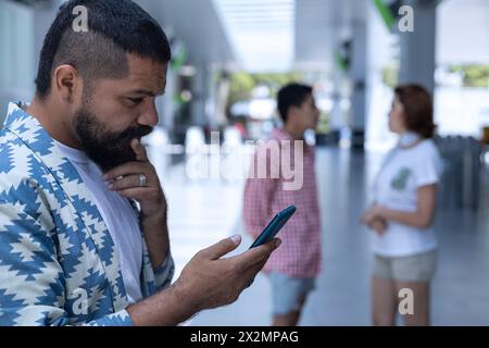 Lateinamerikanischer bärtiger Mann in einem Einkaufszentrum mit seinem Handy. Hintergrund ein paar Freunde unterhalten sich. Kommunikationskonzept Stockfoto