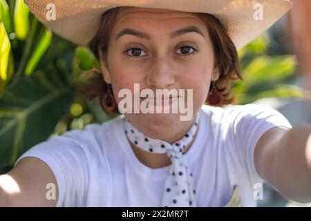 Die junge lateinamerikanische Frau (22) mit Cowboyhut sitzt lächelnd im Park und macht mit ihrem Handy ein Selbstporträt. Konzepttechnologie. Stockfoto