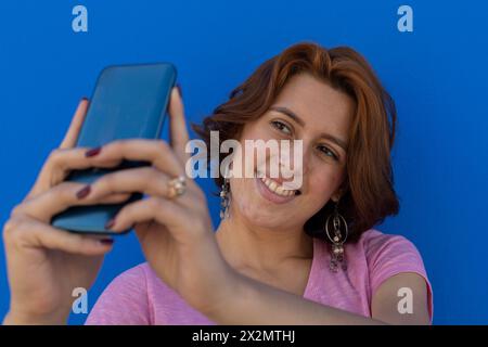 Junge Frau (22) lateinamerikanische Rothaarige, die mit ihrem Handy ein Selbstporträt macht. Blauer Wandhintergrund. Porträt-Konzept. Stockfoto