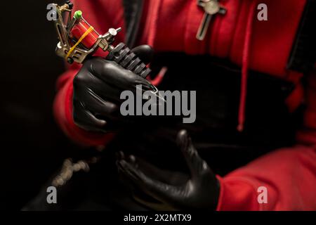 Die Hände des Tätowierers zeigen seine Tätowiermaschine. Er trägt schwarze Handschuhe, roten Pullover und schwarzen Hintergrund. Body-Art-Konzept Stockfoto