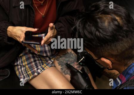 Junge Frau (21), die auf ihrem Smartphone schreibt, während sie von einem professionellen venezolanischen Tätowierer auf ihr linkes Bein tätowiert wurde. Body-Art-Konzept Stockfoto