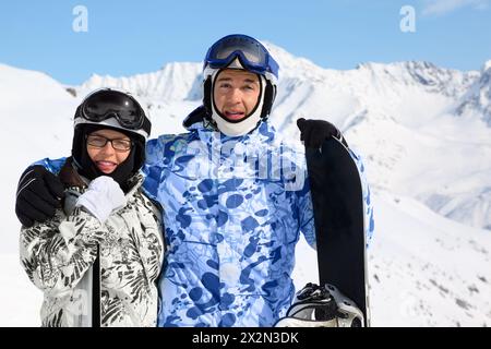 Lächelnde Paare in Sportanzügen und Helmen stehen mit Snowboard und Skiern auf dem Berg. Stockfoto