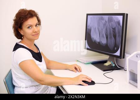 Der Zahnarzt sitzt am Tisch mit einem grossen Kieferröntgenbild auf dem Monitor in der Zahnklinik und schaut in die Kamera Stockfoto