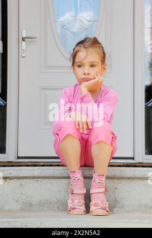 Das kleine traurige Mädchen sitzt auf der Treppe neben der weißen Tür und denkt nach. Stockfoto