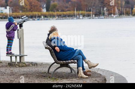 Ein Mann und eine Frau sitzt auf einer Parkbank beisamen und schauen auf den Bodensee. Daneben ein Kind, was mit einem Fernglas spielt. (Konstanz Stockfoto