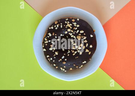 Winzelwinkelaufnahme eines einzelnen Donuts, bedeckt mit Schokolade und bestreuten Nüssen auf einer weißen Platte auf einem einfachen Hintergrund von 3 Farben Stockfoto