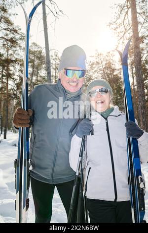 Vertikale mittellange Aufnahme eines fröhlichen aktiven Seniorenpaares in Wintersportbekleidung und Sonnenbrille, die draußen mit Skiern und Stangen posiert Stockfoto