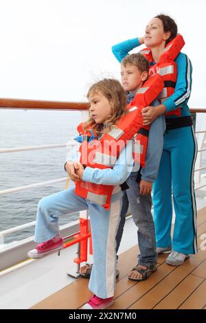 Mutter, Sohn und Tochter in orangefarbenen Schwimmwesten stehen an Deck des Schiffes und blicken auf den Ozean; konzentrieren Sie sich auf das Mädchen; Kinder stehen vor Mama Stockfoto