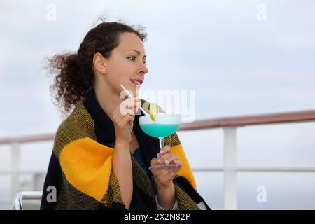 Die lächelnde Frau, in Decke gewickelt, sitzt auf einem Stuhl auf dem Kreuzfahrtschiff-Deck, träumt und hält einen Cocktail Stockfoto