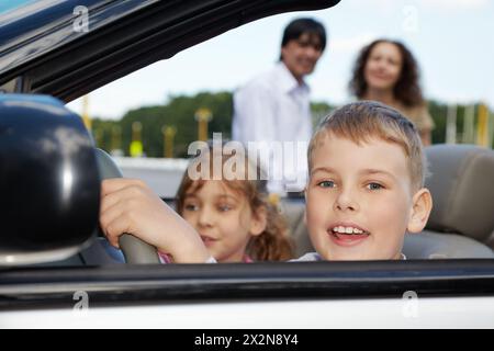 Nahaufnahme des Jungen, der am Lenkrad des Cabriolets sitzt, seine Schwester und Eltern auf Hintergrund nicht im Fokus Stockfoto