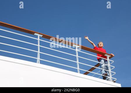 Junge steht am Geländer an Deck des Schiffes, ruft und winkt mit der Hand Stockfoto