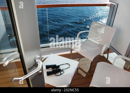 Offene Tür; Glasbalkon mit weißen Stühlen und Tisch mit Fernglas auf dem Kreuzfahrtdeck Stockfoto