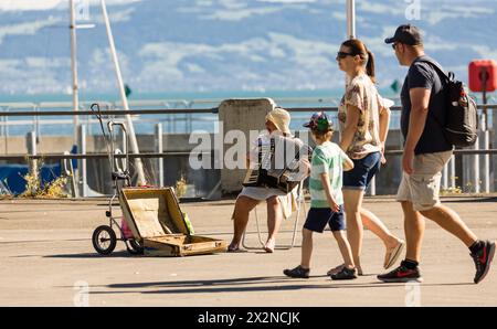 Eine Strassenmusikerin sitzt mit ihrem Handeorgel an der Uferpromenade in Friedrichshafen und spielt Musik. Einige Personen laufen vorbei. (Friedrichs Stockfoto