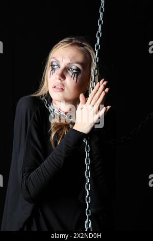 Melancholisches Zombie-Mädchen mit schwarzen Tränen und durchgeschnittener Kehle hält die Kette und blickt auf schwarzen Hintergrund weg. Stockfoto