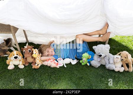 Unter einer großen weißen Decke liegt ein Junge auf dem Gras und legt eine Sammlung von weichen Spielzeugen um ihn herum Stockfoto