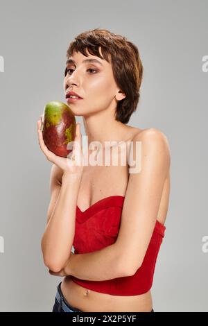 Eine junge Frau in einem roten Oberteil hält eine reife Mango in einer lebendigen Umgebung. Stockfoto