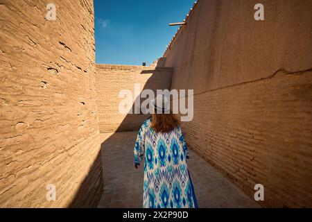 Porträt einer jungen Frau in ethnischer Kleidung mit blauem Ornament und Hut, die an der Stadtmauer in Itchan Kala in der alten Stadt Chiwa in Usbekistan spaziert. Stockfoto