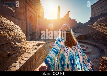 Frau in ethnischer Kleidung im Innenhof in der Nähe der alten Madrasah, die Mann von Hand an der alten Stadtmauer in Chiwa in Usbekistan hält. Stockfoto