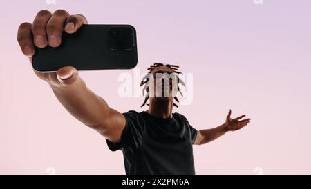 Stilvoller junger Mann mit Dreadlocks posiert selbstbewusst in einem Studio. Mit seinem Handy und seiner Selfie-Kamera erfasst er einen Vlog für seine Stockfoto