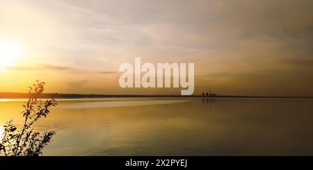 Die Sonne bei Sonnenuntergang über einem Zweig eines großen Sees und Fabriken am Horizont Stockfoto