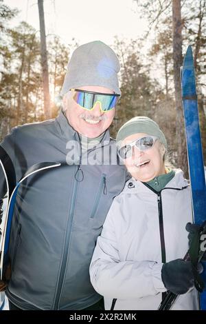 Vertikales mittleres Porträt eines glücklichen älteren Mannes und einer Frau in Wintersportbekleidung mit Sonnenbrille, die draußen steht und in die Kamera lächelt Stockfoto