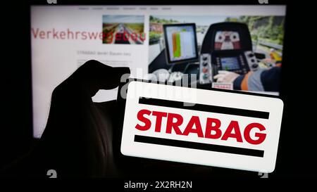 Person, die ein Mobiltelefon mit dem Logo des österreichischen Bauunternehmens Strabag SE vor der Business-Webseite hält. Schwerpunkt auf dem Display des Telefons. Stockfoto