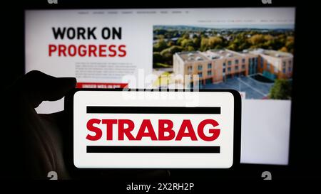 Person mit Smartphone mit Logo des österreichischen Bauunternehmens Strabag SE vor der Website. Schwerpunkt auf dem Display des Telefons. Stockfoto