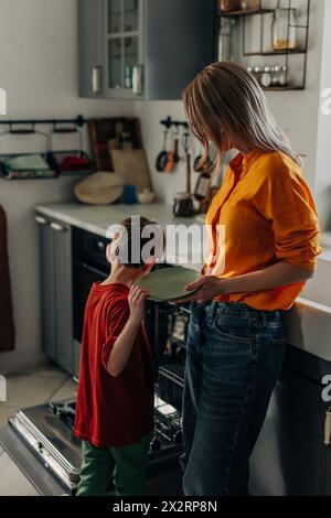 Sohn Entladeplatte aus dem Geschirrspüler mit Mutter in der Küche zu Hause Stockfoto