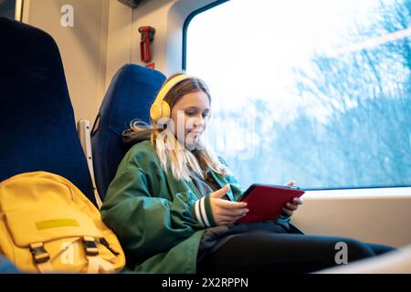 Mädchen mit schnurlosen Kopfhörern, die einen Tablet-PC im Zug in der Nähe des Fensters verwenden Stockfoto