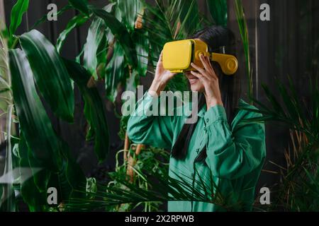 Glückliche Geschäftsfrau mit gelbem Virtual-Reality-Simulator in der Nähe von Pflanzen Stockfoto