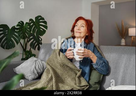 Eine ältere Frau sitzt entspannt unter der Decke auf dem Sofa und trinkt zu Hause Tee Stockfoto