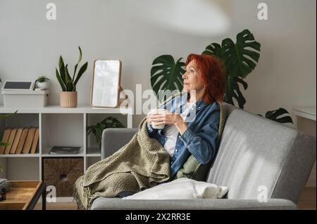 Eine nachdenkliche Seniorin sitzt entspannt unter der Decke auf dem Sofa und trinkt zu Hause Tee Stockfoto