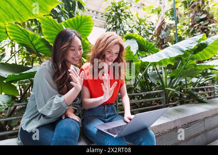 Freunde, die bei Videogesprächen durch den Laptop winken und auf der Bank in der Nähe von Pflanzen sitzen Stockfoto