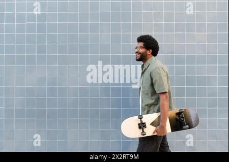 Glücklicher Mann, der mit Skateboard vor der gefliesten Wand läuft Stockfoto