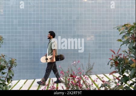 Junger Mann, der mit Skateboard vor einer blau gekachelten Wand läuft Stockfoto