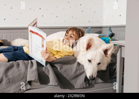 Junge, der auf dem Bett liegt und ein Buch liest, mit weißem Schweizer Hirtenhund Stockfoto
