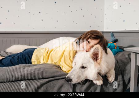 Lächelnder Junge, der mit weißem Schweizer Hirtenhund zu Hause auf dem Bett liegt Stockfoto