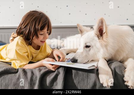 Ein lächelnder Junge liest Buch mit weißem Schweizer Schäferhund auf dem Bett Stockfoto