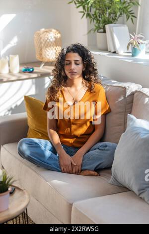 Junge Frau, die zu Hause mit geschlossenen Augen auf dem Sofa meditiert Stockfoto