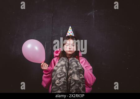 Trauriges Geburtstagskind in Partyhut mit rosa Ballon vor schwarzem Hintergrund Stockfoto