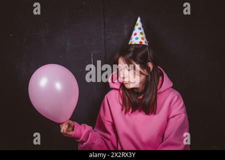 Trauriges Geburtstagsmädchen in Partyhut, das auf rosa Ballon vor schwarzem Hintergrund blickt Stockfoto