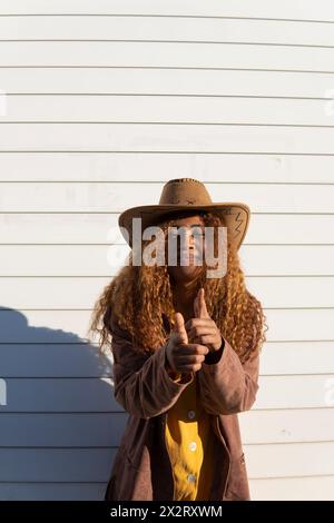 Lächelnde junge Frau mit lockigen Haaren, die Cowboyhut trägt und an sonnigem Tag Gesten macht Stockfoto
