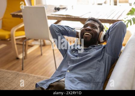 Lächelnde, nicht binäre Person, die schnurlose Kopfhörer trägt und zu Hause Musik genießt Stockfoto