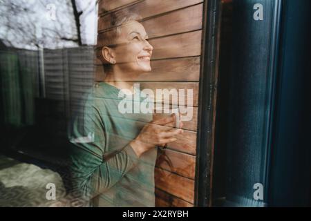Lächelnde Frau, die Kaffeetasse hält und neben dem Glasfenster steht Stockfoto