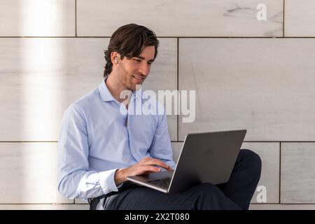 Lächelnder junger Geschäftsmann, der vor der Wand einen Laptop benutzt Stockfoto