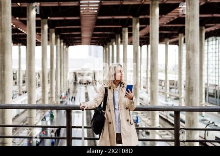 Eine nachdenkliche Frau, die ihr Smartphone hält und am Bahnhof in der Nähe des Geländes steht Stockfoto