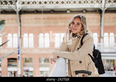 Lächelnde Frau, die sich am Ellenbogen in der Nähe des Geländes am Bahnhof lehnt Stockfoto