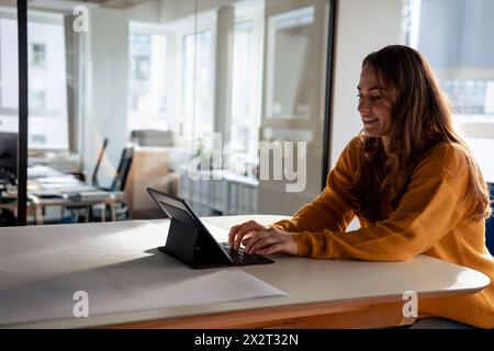 Lächelnde Geschäftsfrau, die auf der Tastatur schreibt und im Büro einen Tablet-PC verwendet Stockfoto